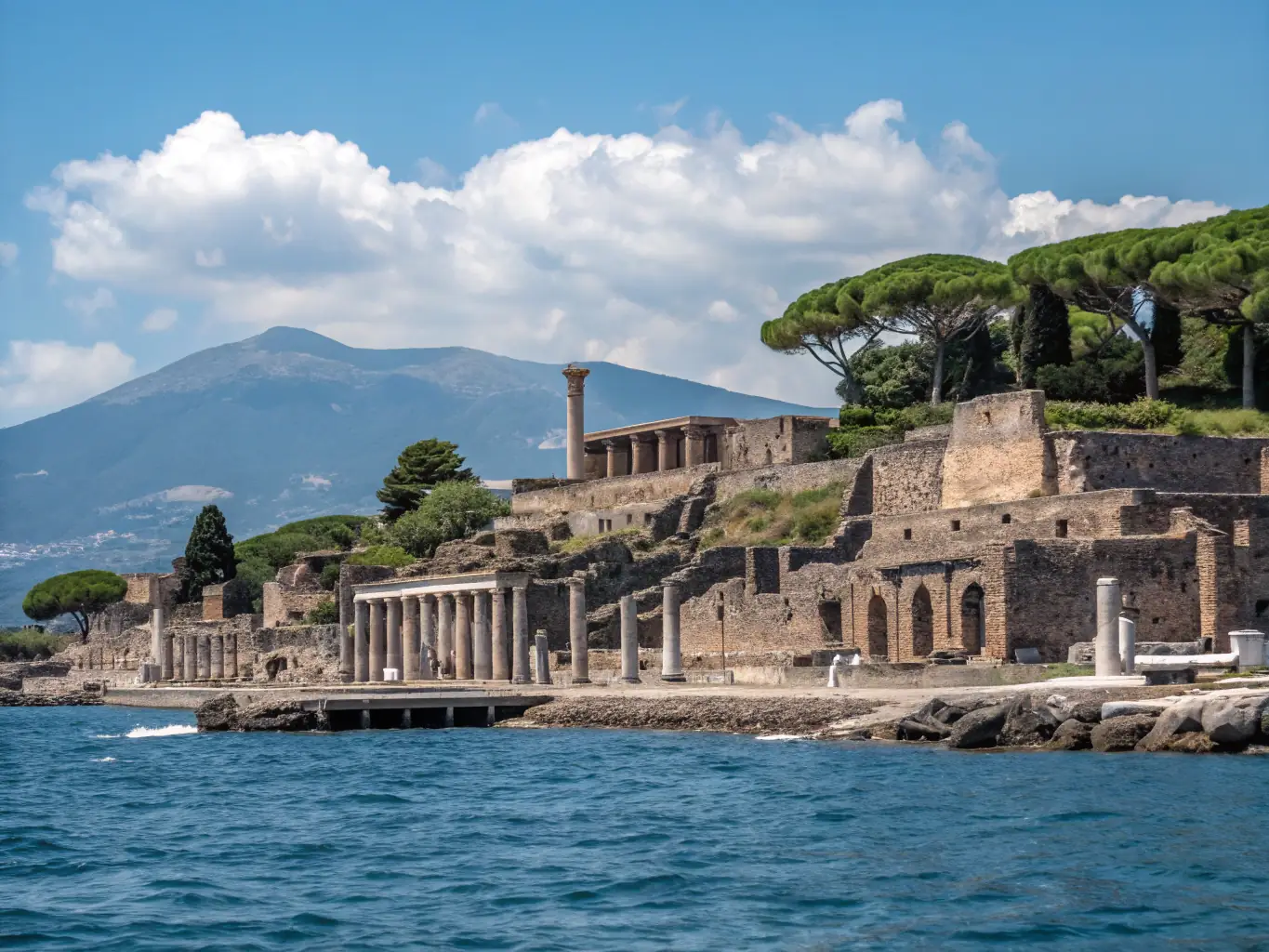 An aerial view of the ancient ruins of Pompeii, showcasing the well-preserved structures and the vastness of the historical site. Tourists can be seen exploring the area, highlighting its popularity as a day trip destination.
